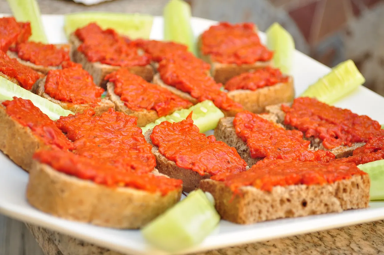 Slices of brown bread thickly spread with bright red ajvar on a white plate with cucumber wedges
