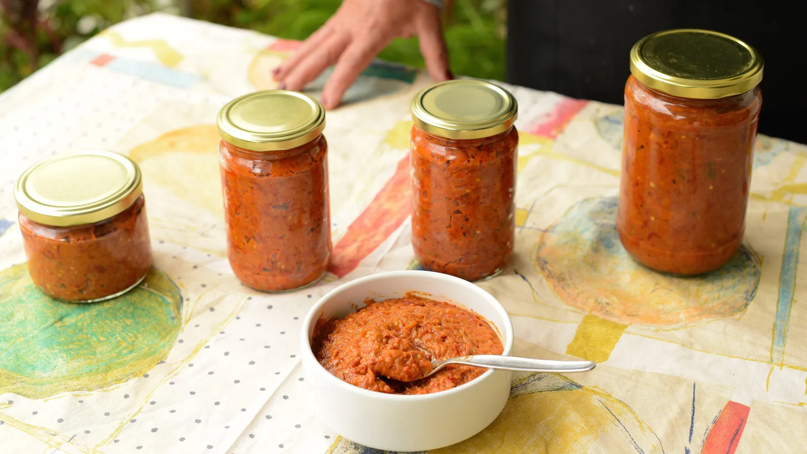 Four glass jars of homemade ajvar and a bowl of the red pepper spread on a garden table