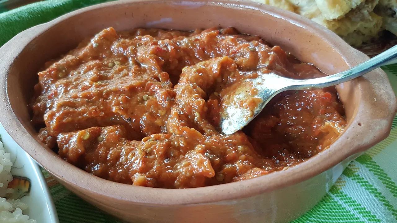 A clay dish of coarse homemade ajvar with aubergine, a spoon resting in it, on a green striped cloth