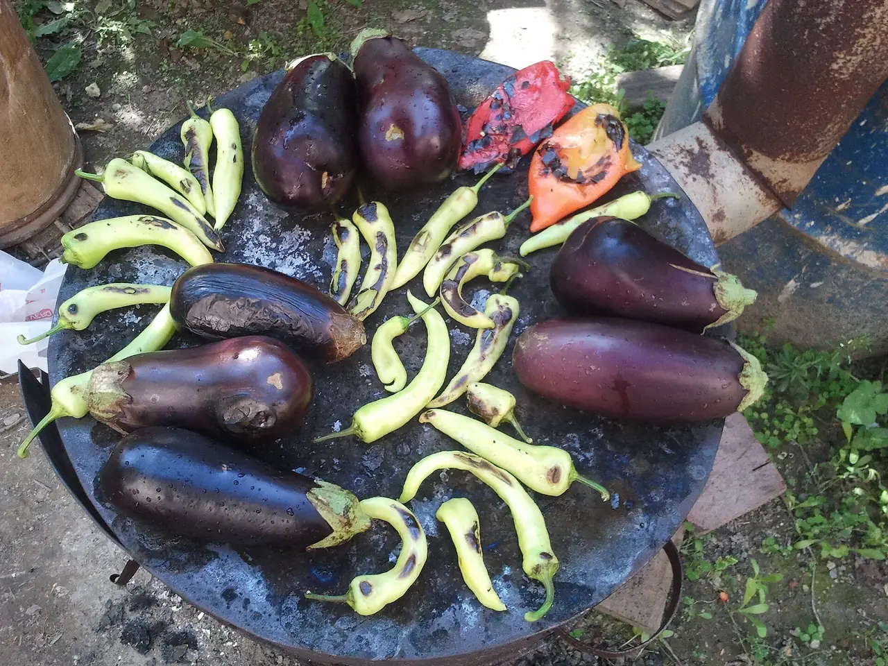 Aubergines and pale green peppers blistering on a round black iron roasting plate set over a fire outdoors