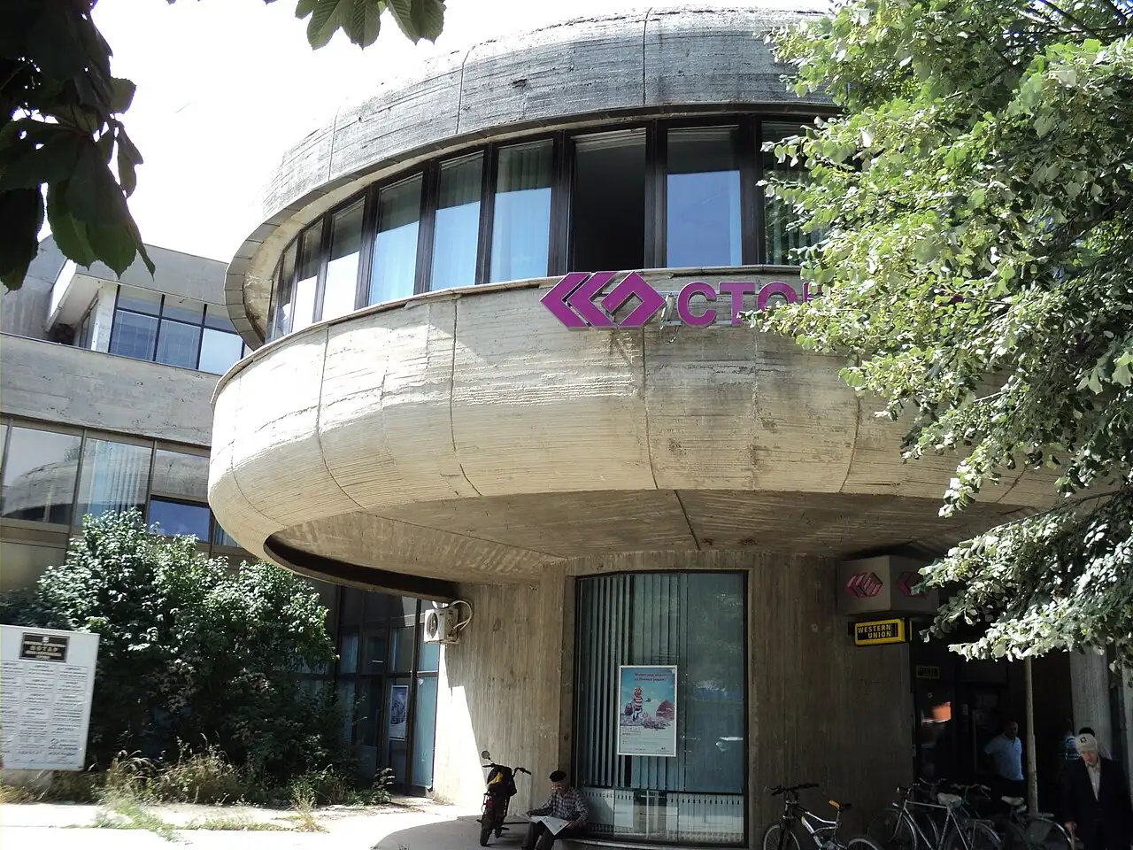 A round brutalist-style Stopanska Banka branch building with a Western Union sign, bicycles parked outside