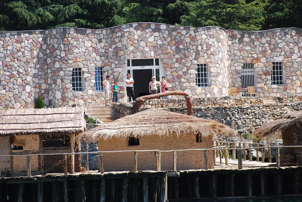 The stone museum reception building of the Bay of Bones with visitors standing on the steps above the thatched huts