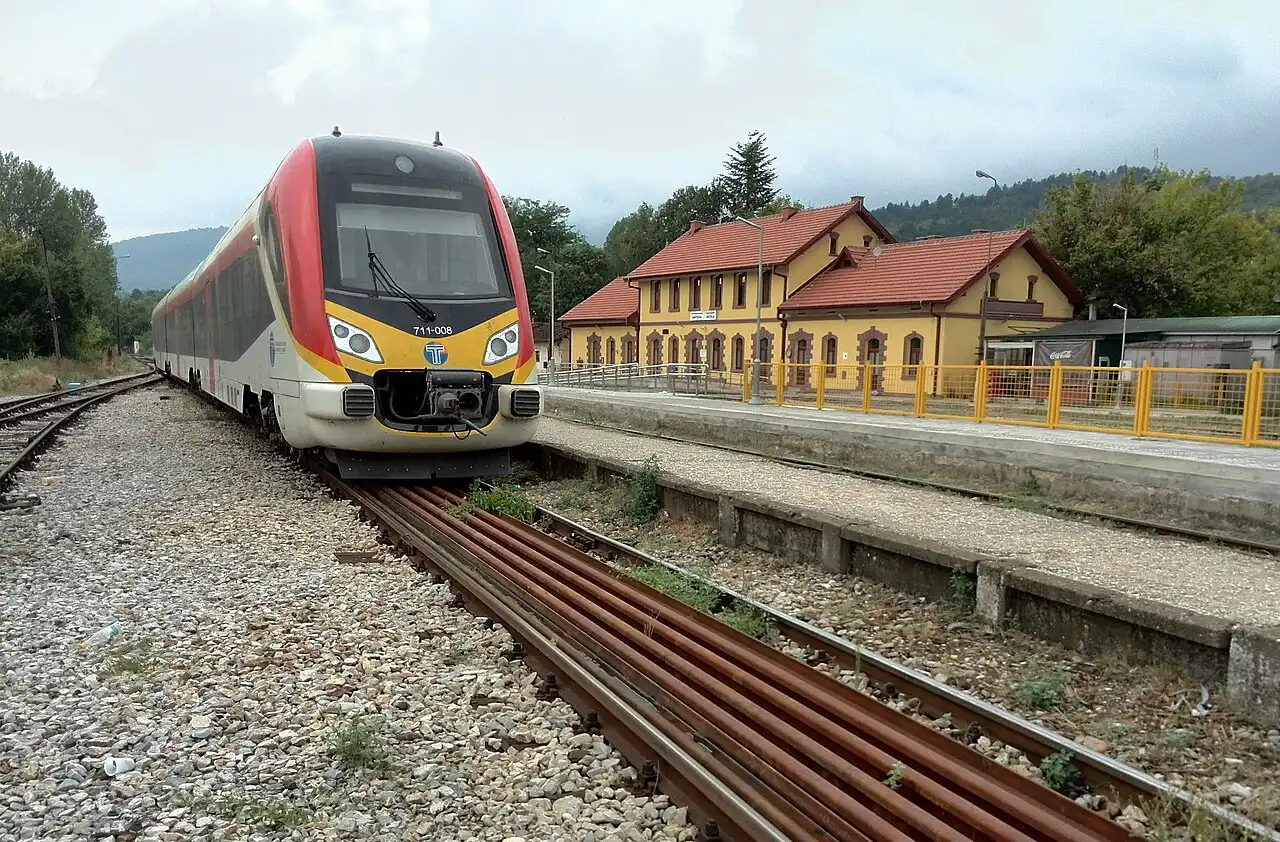 A modern red-and-grey diesel railcar of Macedonian Railways Transport stopped at Bitola's historic yellow railway station building