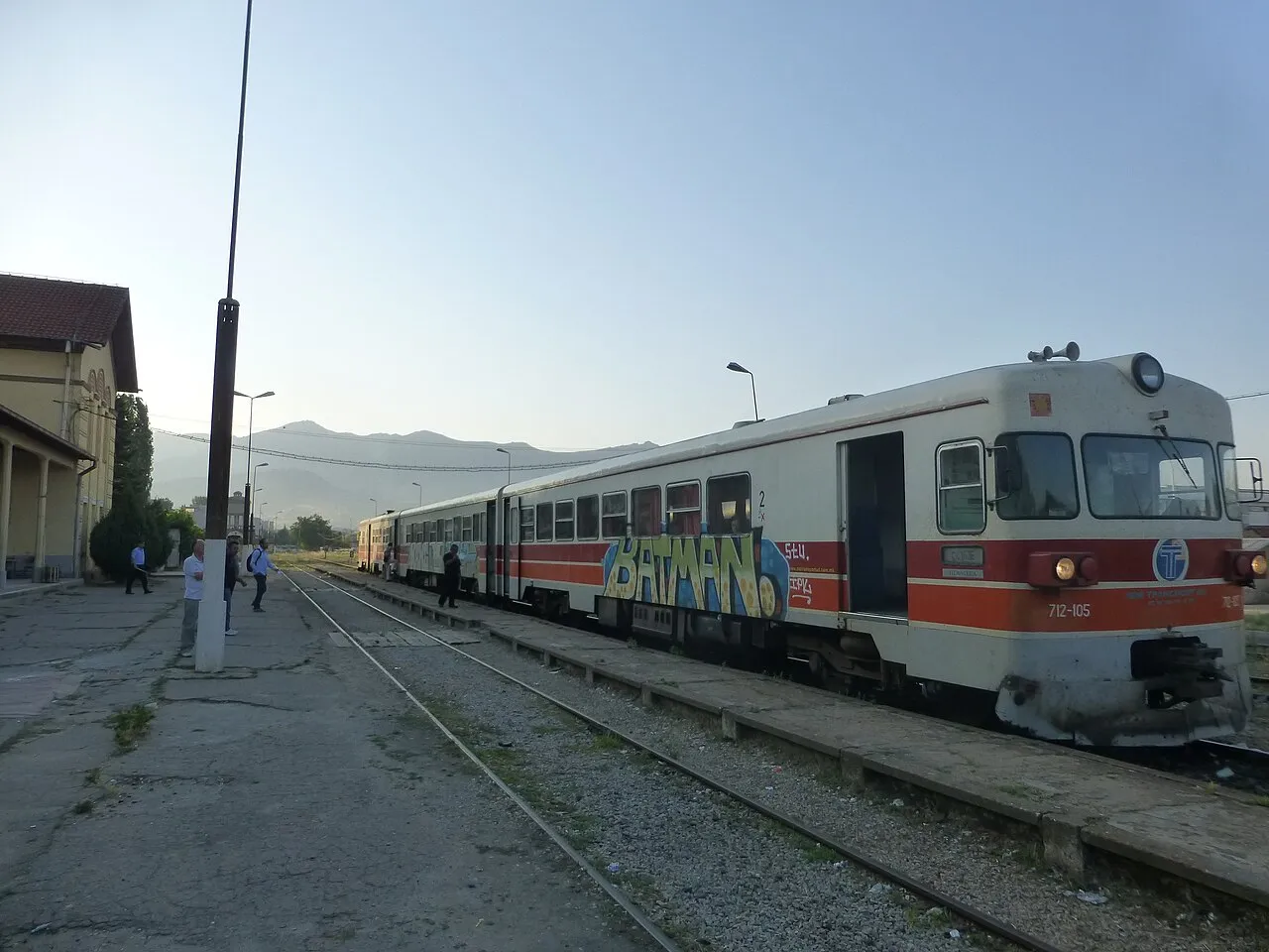 A red-and-grey Macedonian Railways Transport diesel railcar stopped at Prilep's own station building, with passengers on the platform and hills behind