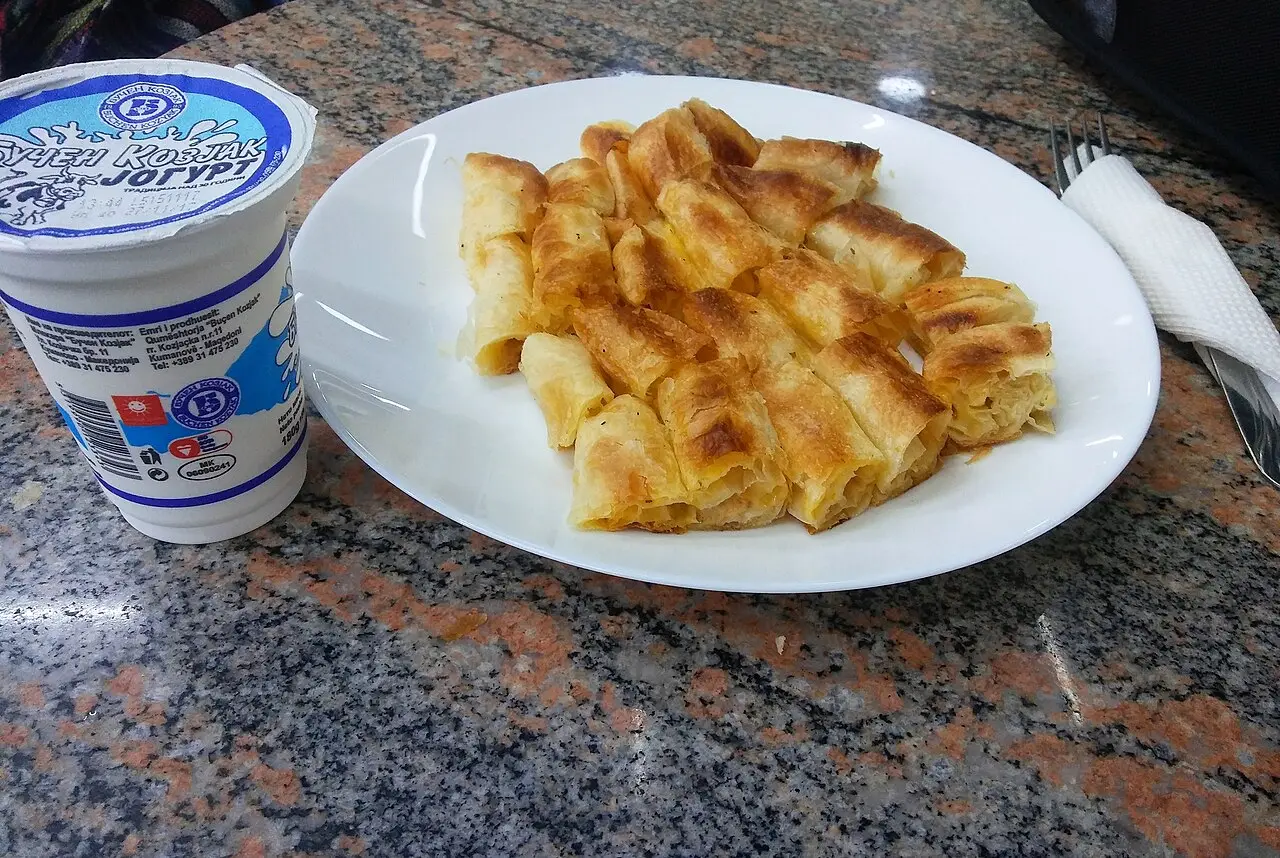 A plate of burek pieces beside a cup of drinking yoghurt on a granite counter