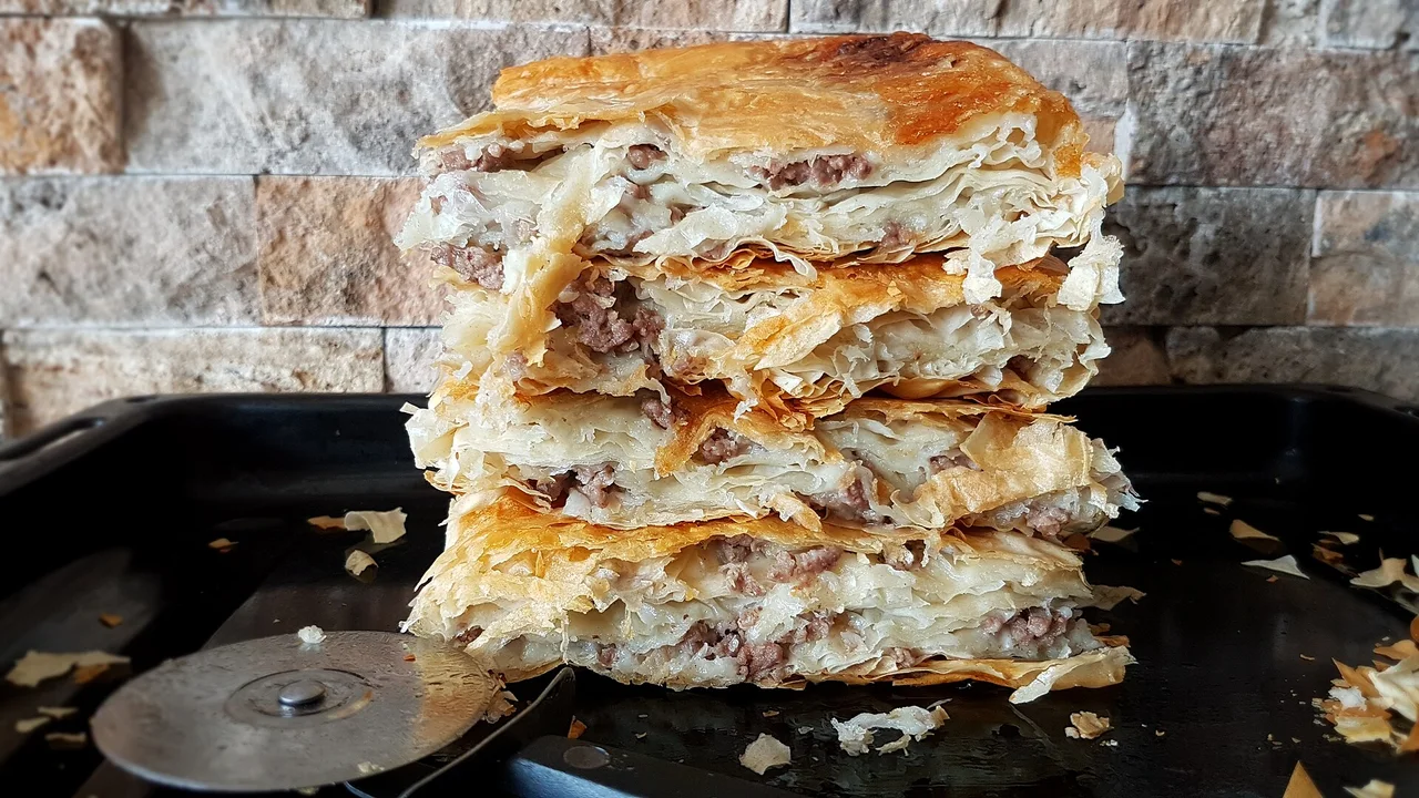 A tall stack of cut burek showing thin sheets of pastry around minced meat and onion, with a pizza wheel in the tray