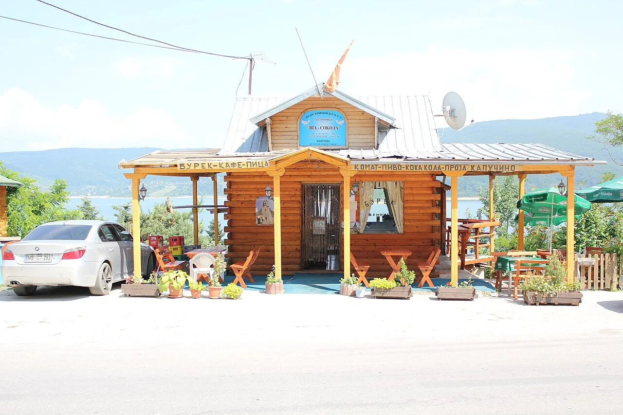 A log-cabin burek, cafe and pizza stand with a terrace at Mavrovi Anovi, with Lake Mavrovo behind it