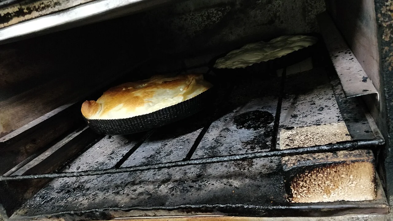 Two round burek tins in a bakery oven, one baked golden and the other still pale