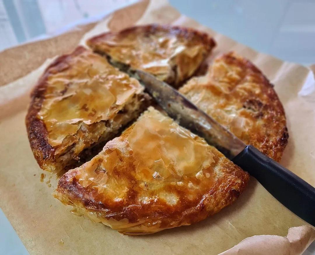 A round meat burek cut into four wedges on baking paper, with a knife lying across it