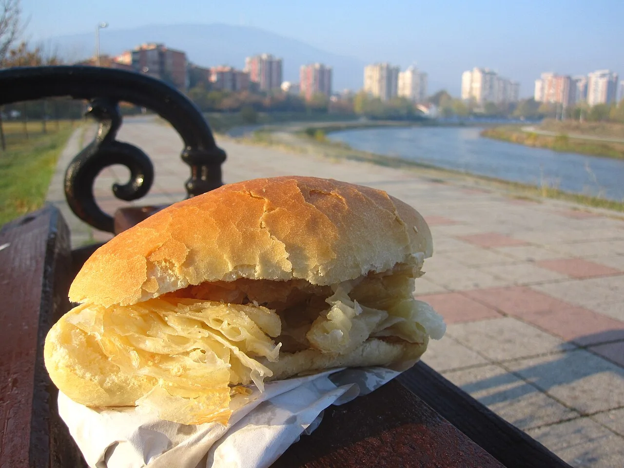 A simit-pogača bun split open and stuffed with shredded burek pastry, resting on a park bench above a river