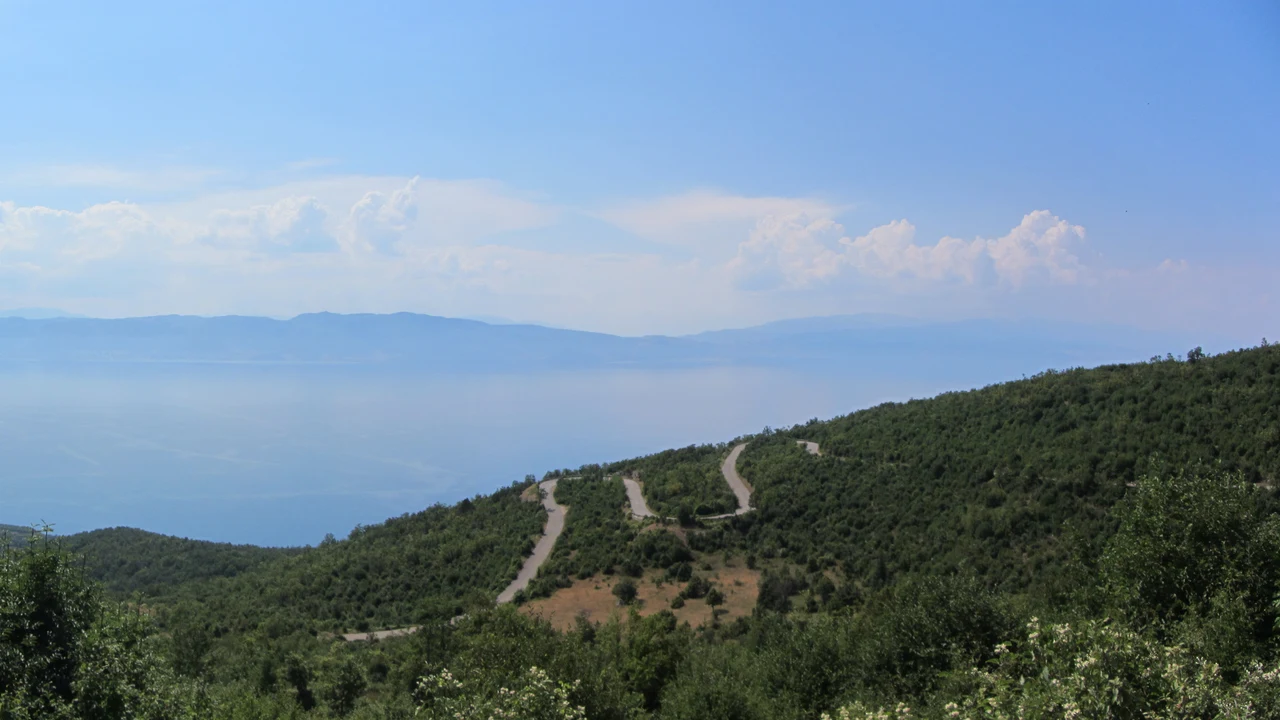 The Galicica mountain road switchbacking down wooded slopes above the hazy blue expanse of Lake Ohrid