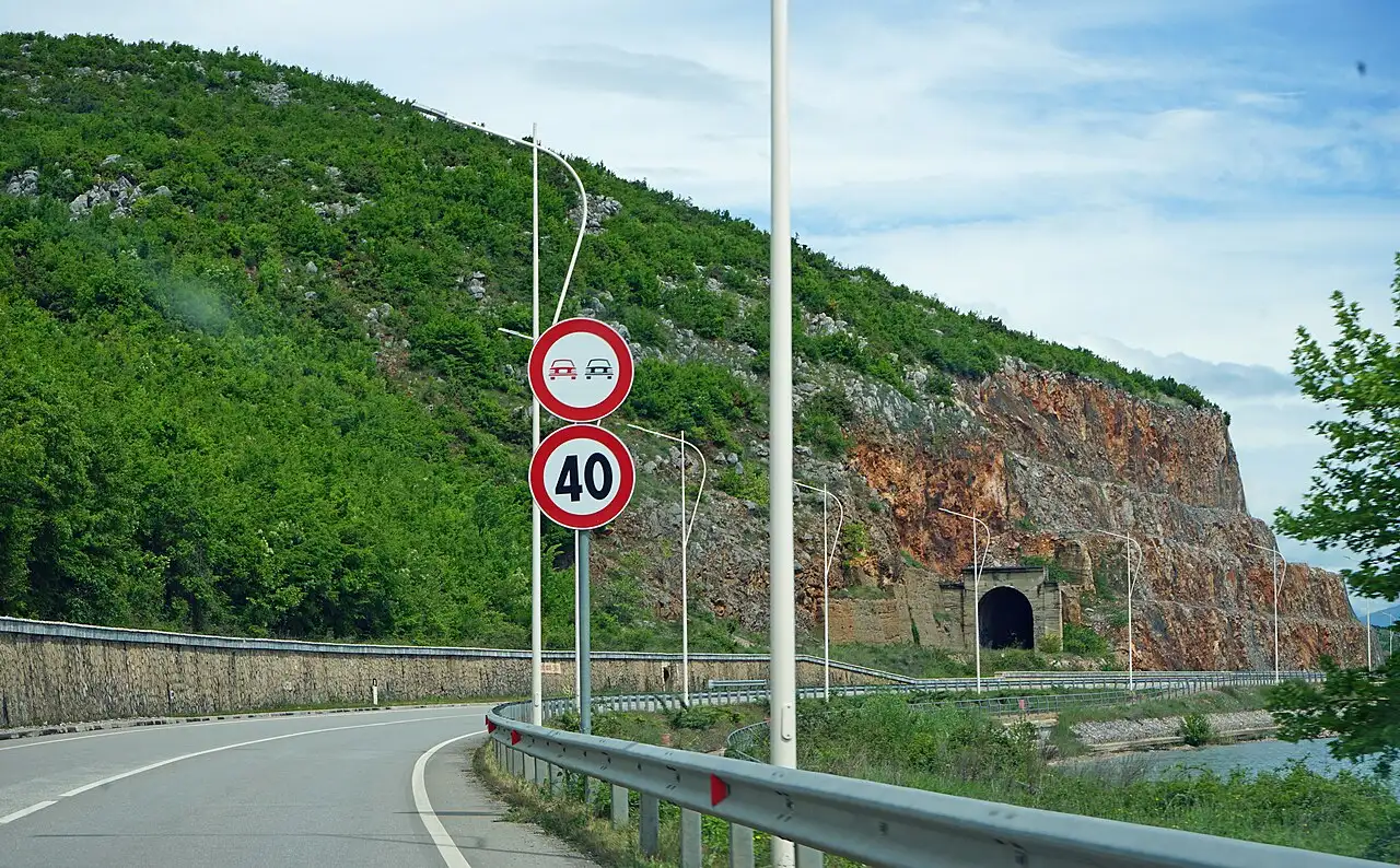 A lakeside road on the Albanian shore of Lake Ohrid with a 40 km per hour sign, a no-overtaking sign and an old railway tunnel portal