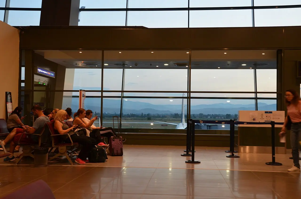 Passengers waiting by the gates at Skopje airport at dusk with an aircraft outside the window