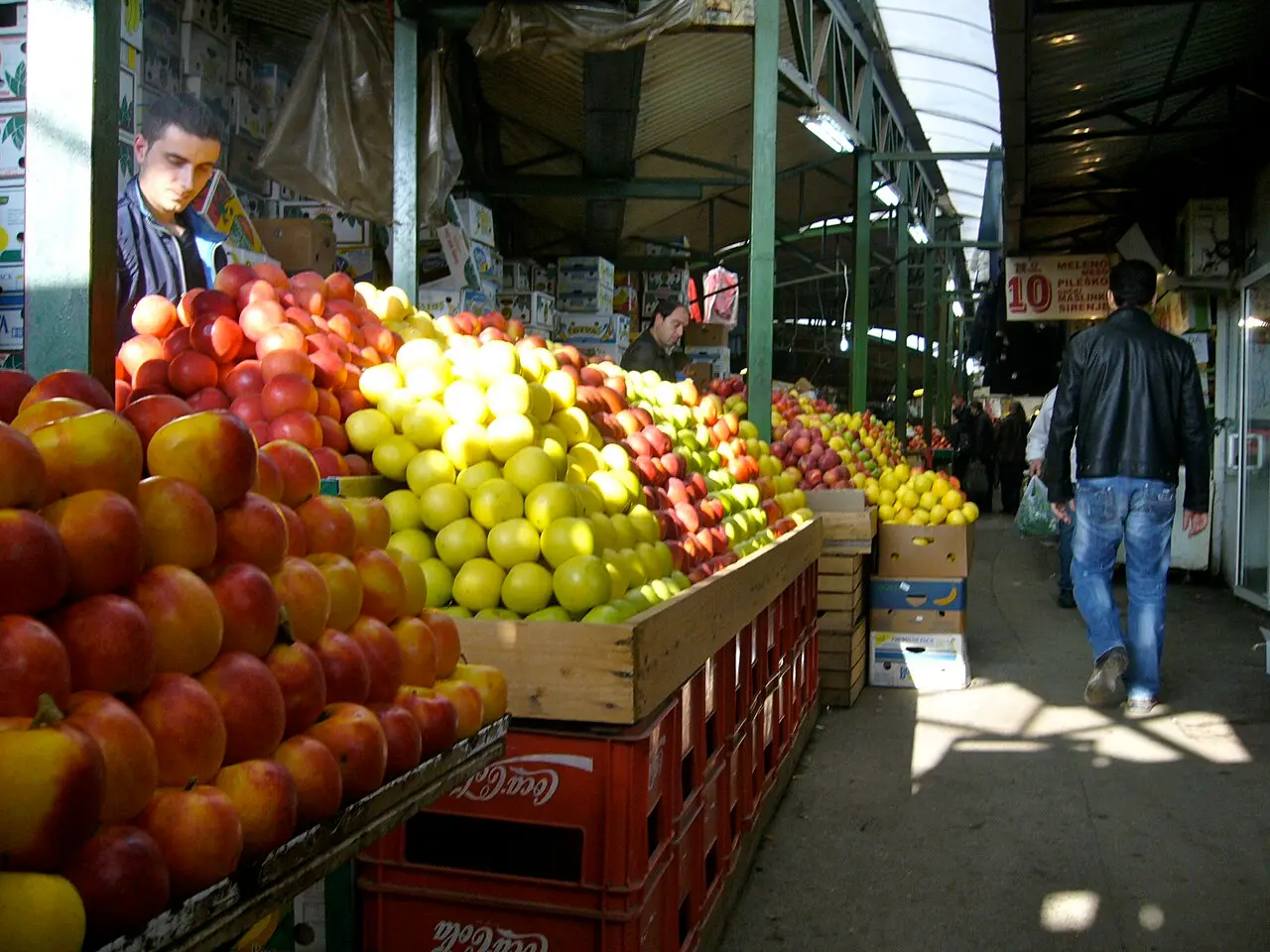 Stacked pyramids of apples and nectarines at a covered green market in Skopje, with a shopper walking past