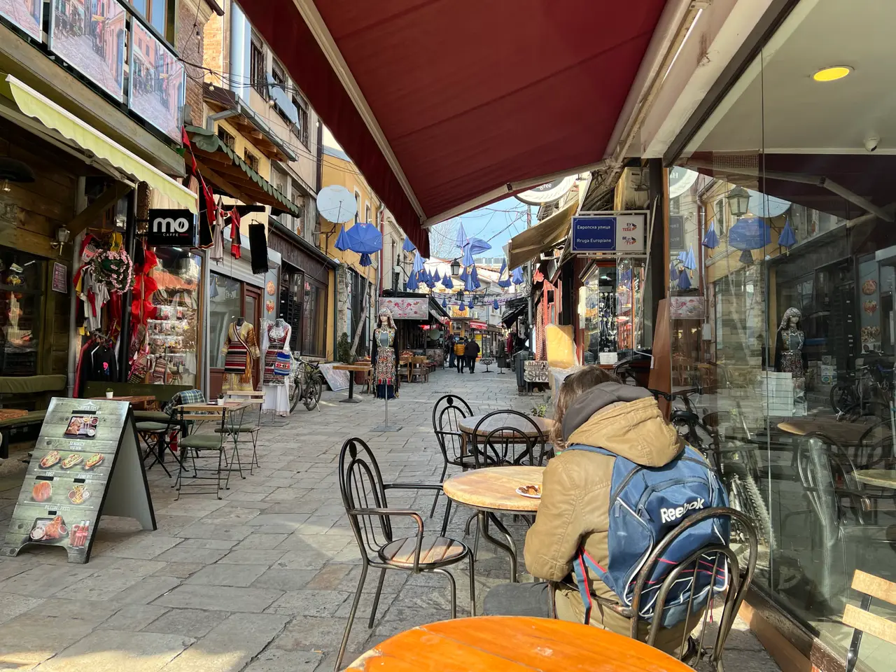 A person sitting with a backpack at an outdoor café table under an awning on a paved lane in Skopje Old Bazaar