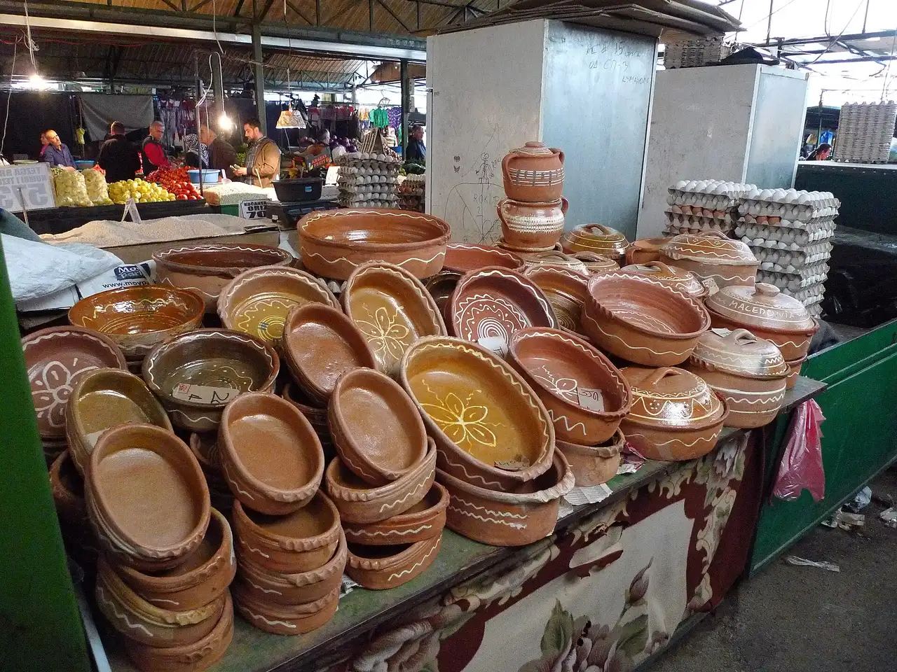 Piles of glazed terracotta baking dishes and lidded pots on a market stall, with egg trays and vegetable counters behind