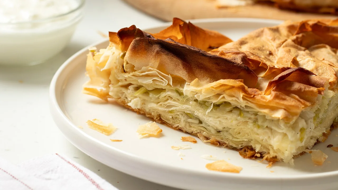 A slice of homemade leek burek on a white plate with a bowl of yogurt behind it, the thin pastry layers showing at the cut edge