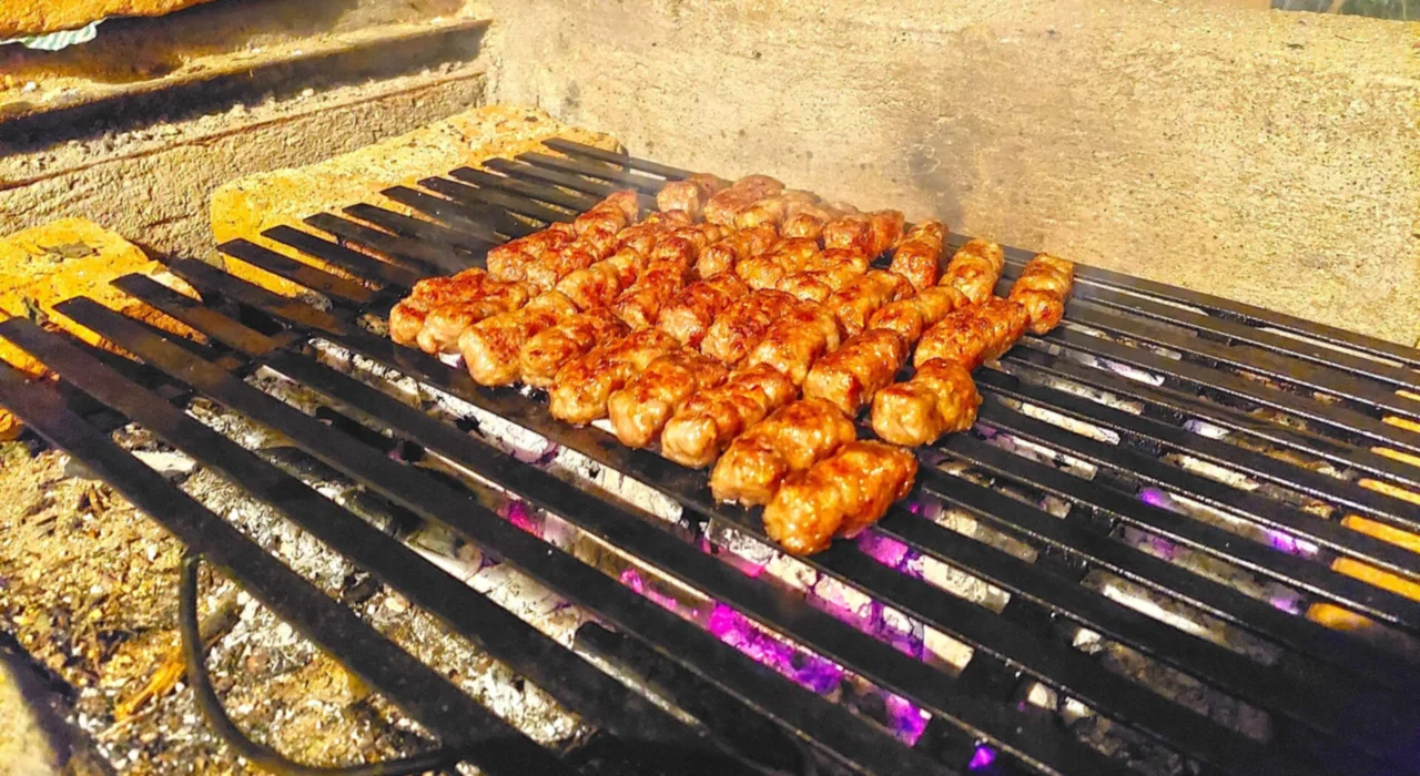 Rows of kebapi, small finger-shaped grilled minced-meat sausages, browning on an iron grill over glowing charcoal
