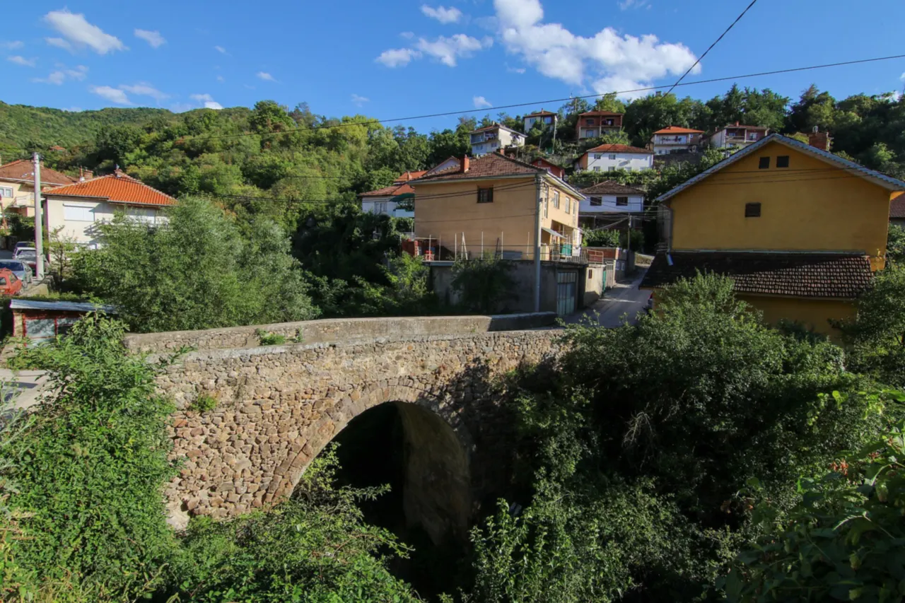Old stone arch bridge in Kratovo with yellow and white houses and wooded hills behind it under a blue sky