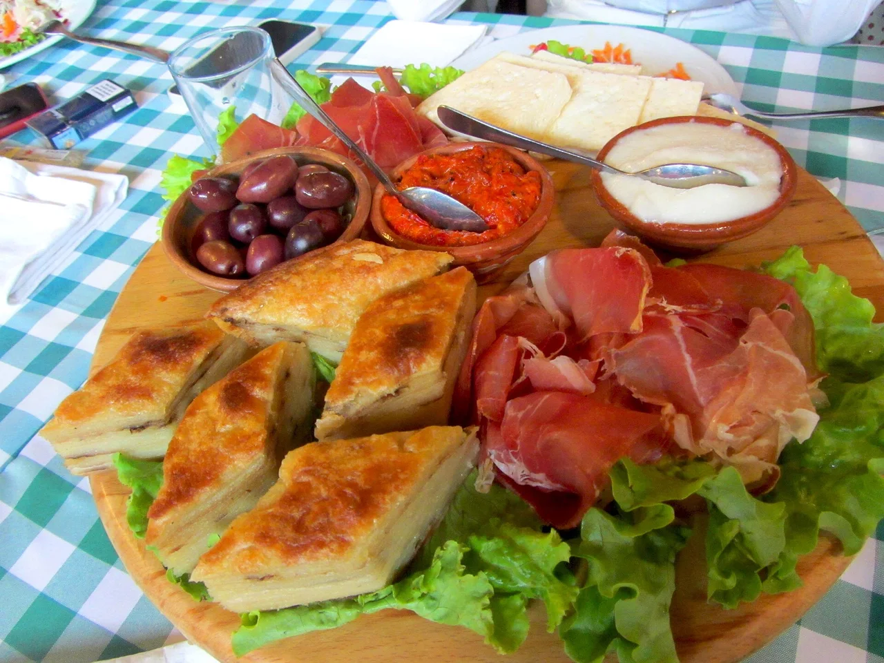 Wooden platter of cured ham, golden pastry squares, white cheese, black olives, red pepper relish and a yogurt dip on a checked tablecloth