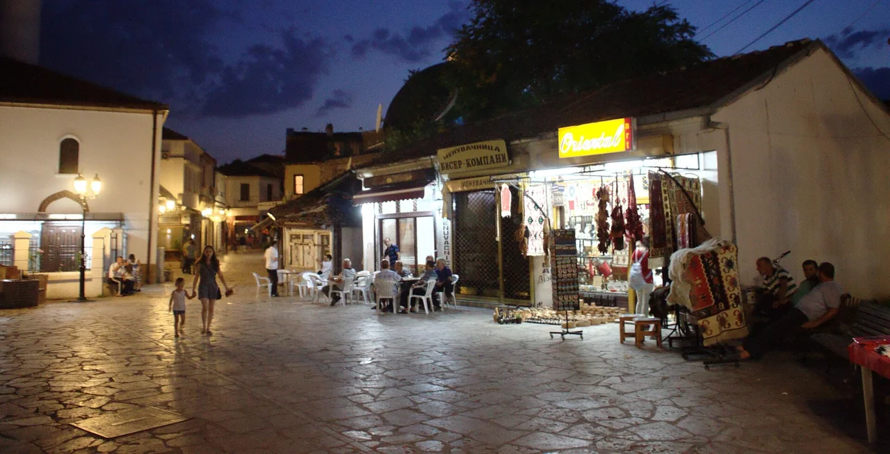Stone-paved square in Skopje's Old Bazaar at dusk, with people at white café tables and a carpet shop still lit beside them