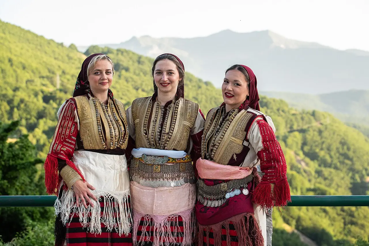 Three women in heavy embroidered Galichnik folk costume with coin belts and red aprons, mountains behind