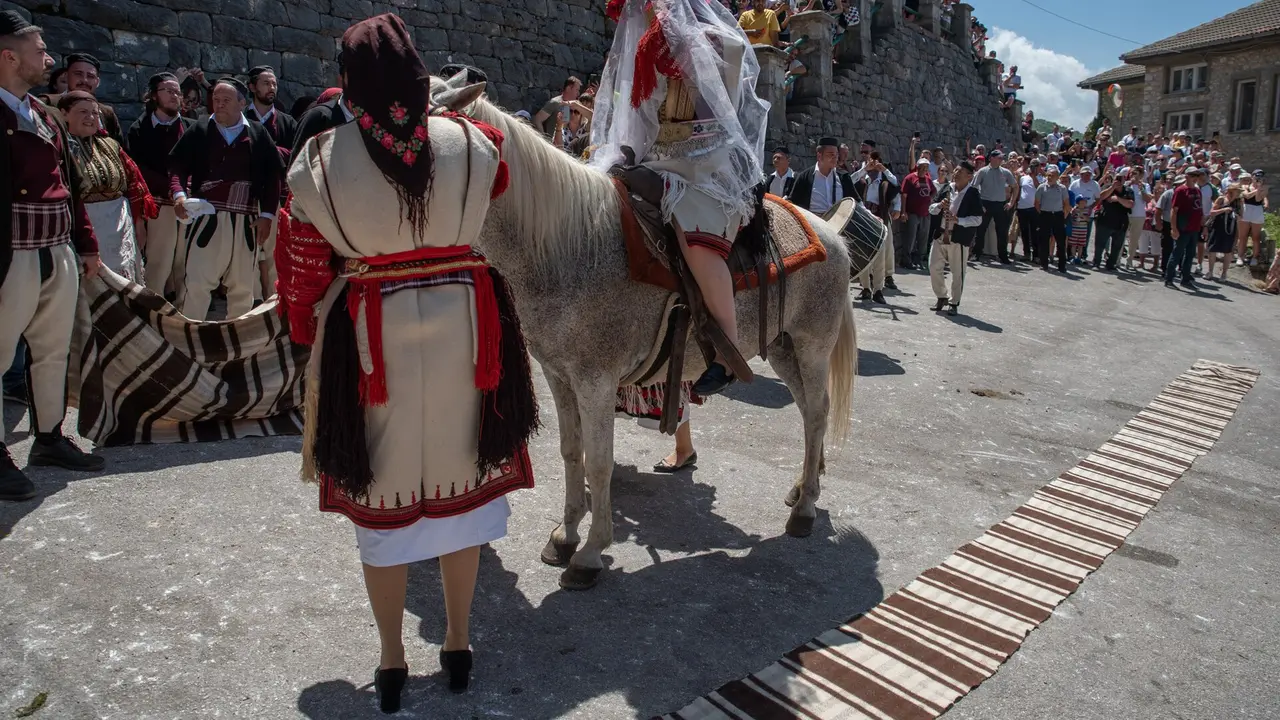 A bride in traditional dress leads a white horse toward the church as costumed villagers and a drummer look on at the Galichnik Wedding