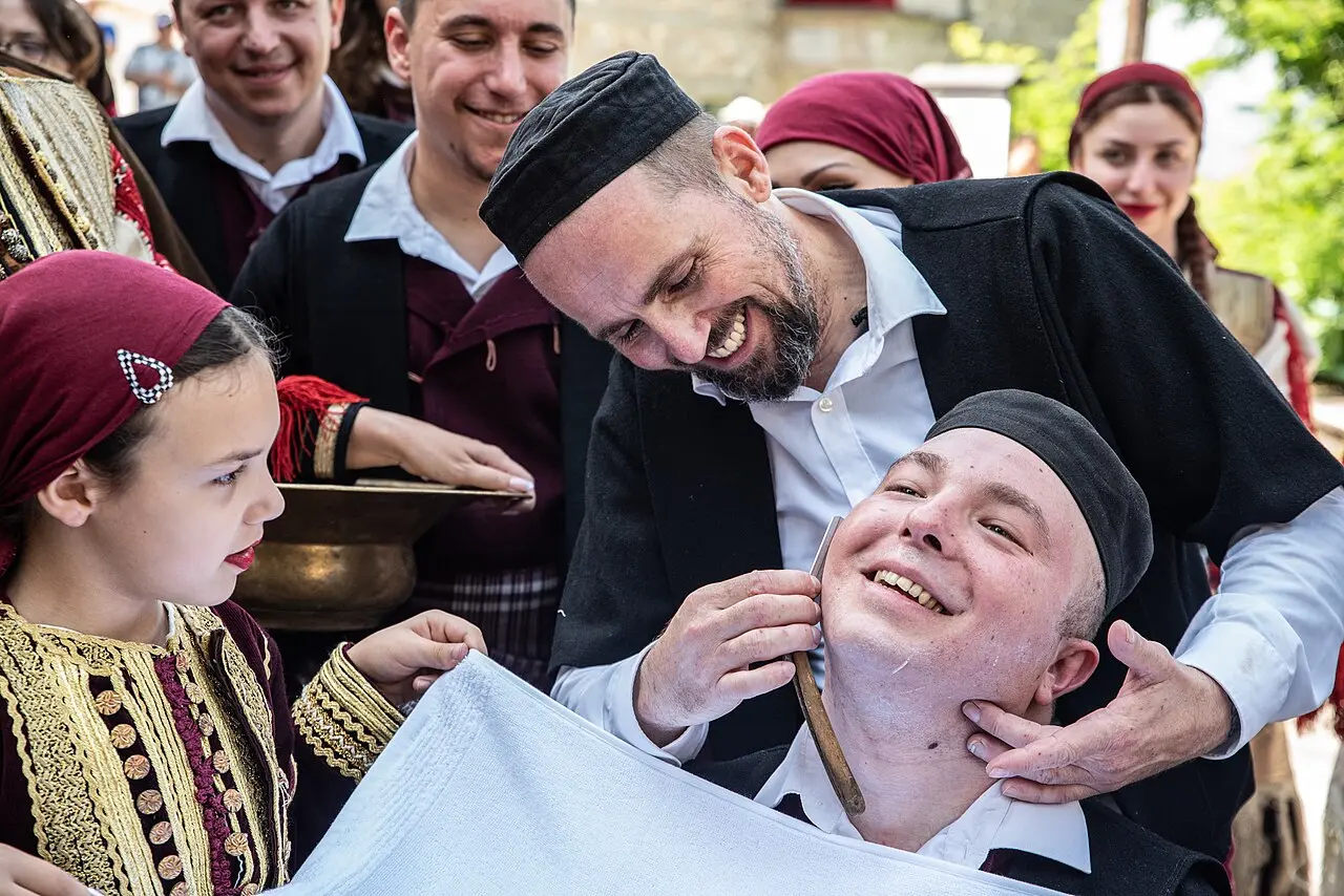 A man shaves the smiling groom with a straight razor while costumed guests, including a girl in traditional dress, look on