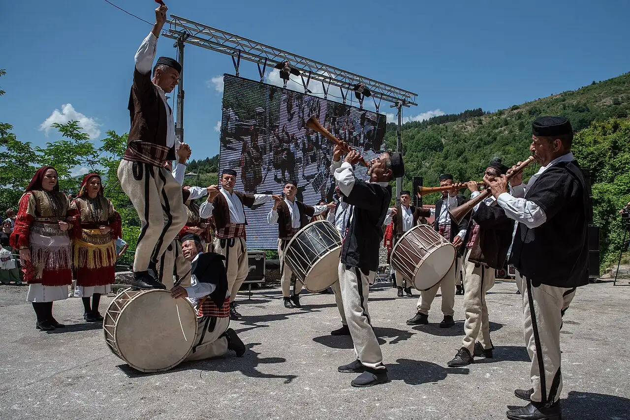 Men in white folk costume dance the Teshkoto in motion beside a large drum