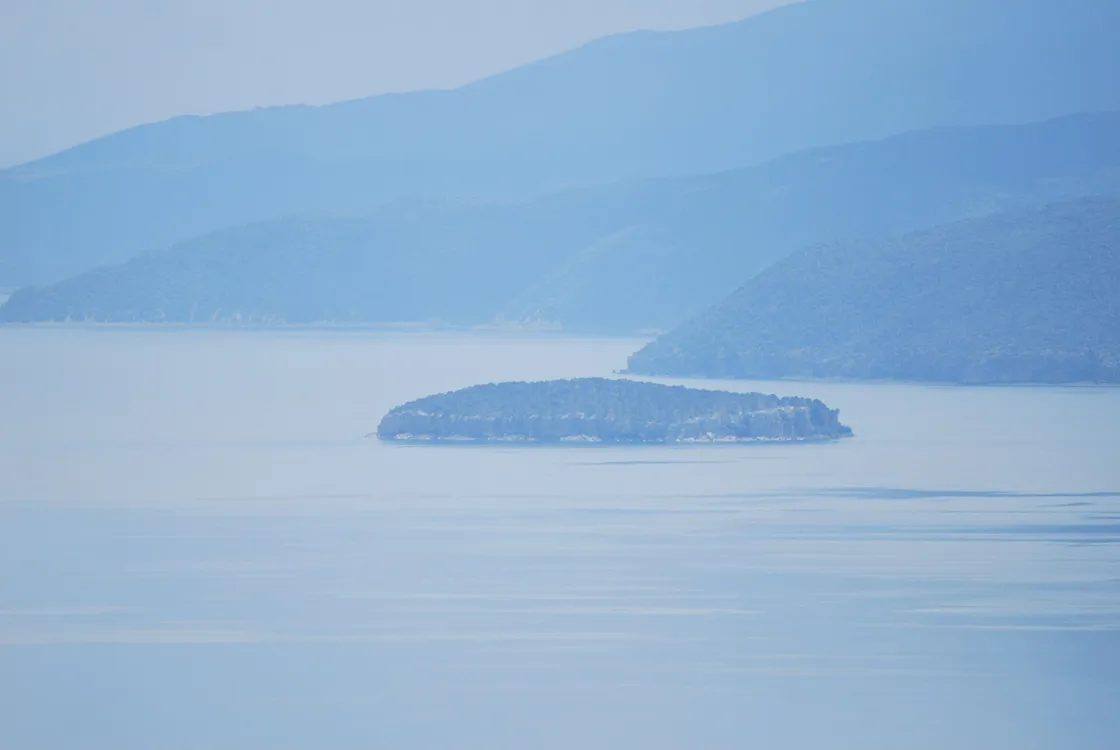 The wooded islet of Golem Grad floating on the hazy blue water of Lake Prespa with hills behind