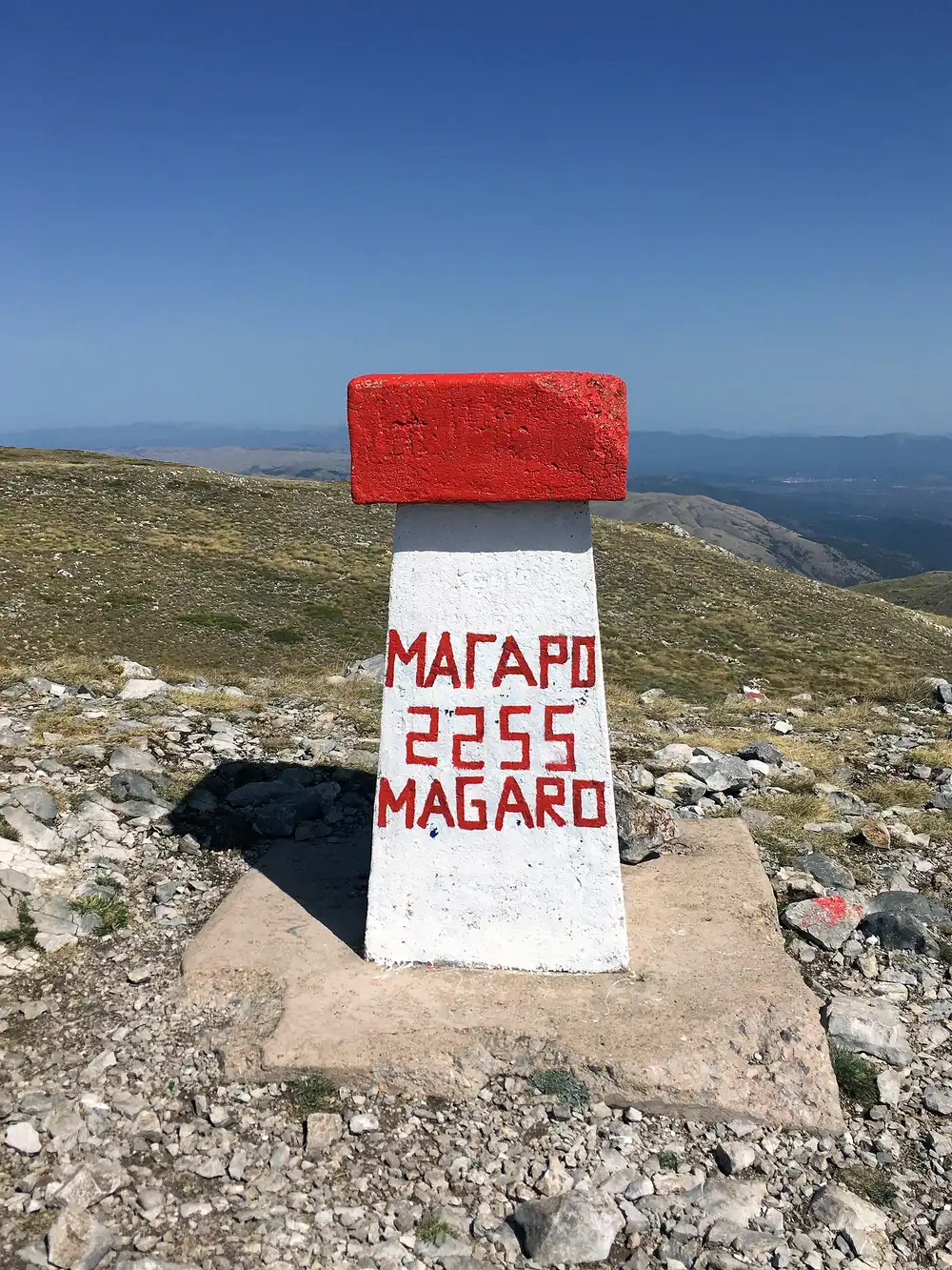 The white and red painted summit marker of Magaro reading 2255 on the bare grassy top of Galičica
