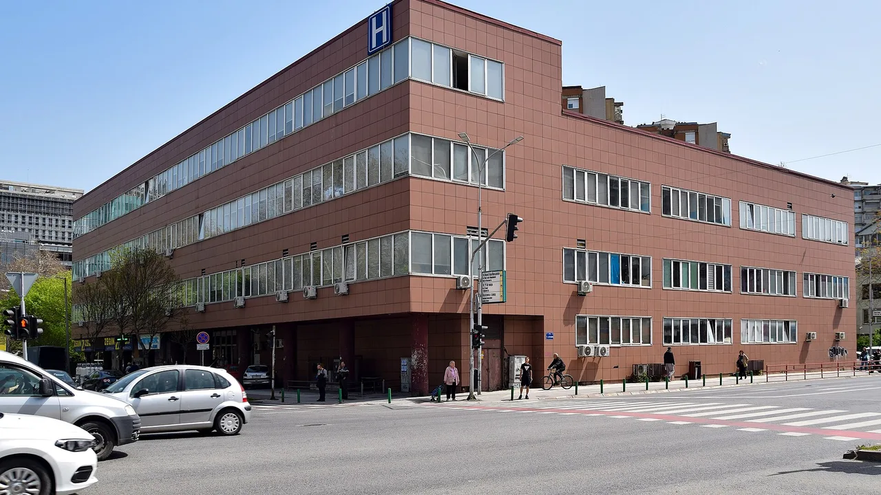 Public city hospital building in Skopje with a blue H sign on the corner, a parked taxi and traffic on the street