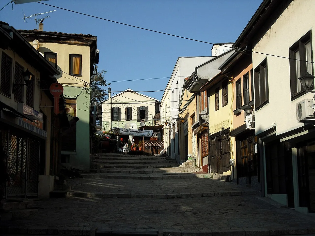 A lane in Skopje Old Bazaar lined with small shops and cafes under a strip of blue sky