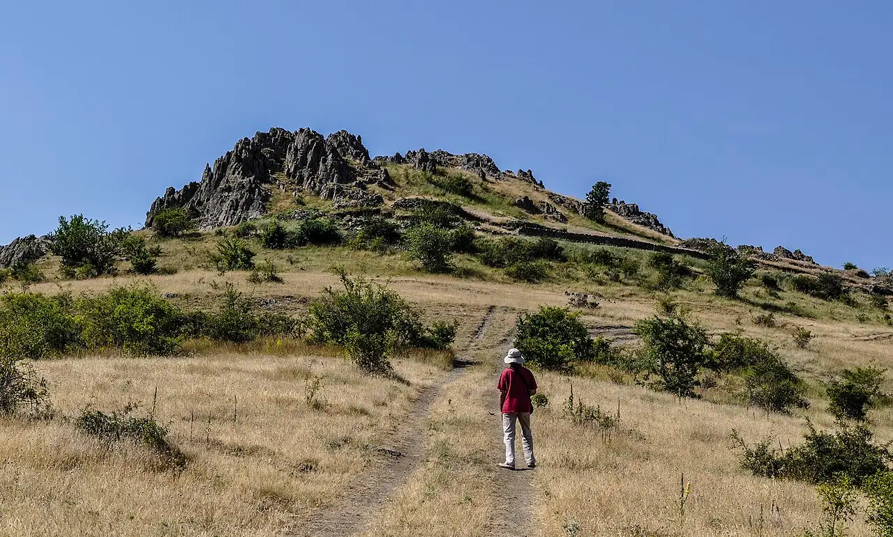 A walker on the dirt path leading up the grassy slope to the rocky summit of Kokino