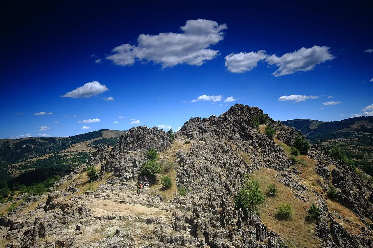 The rocky andesite crest of Kokino rising above dry grass under a blue sky with clouds