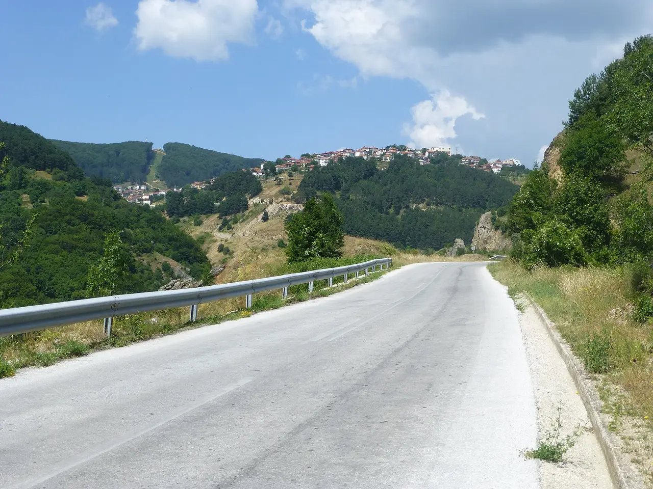 The winding mountain road up to Kruševo, its red-and-white houses spread along a wooded ridge ahead under summer clouds