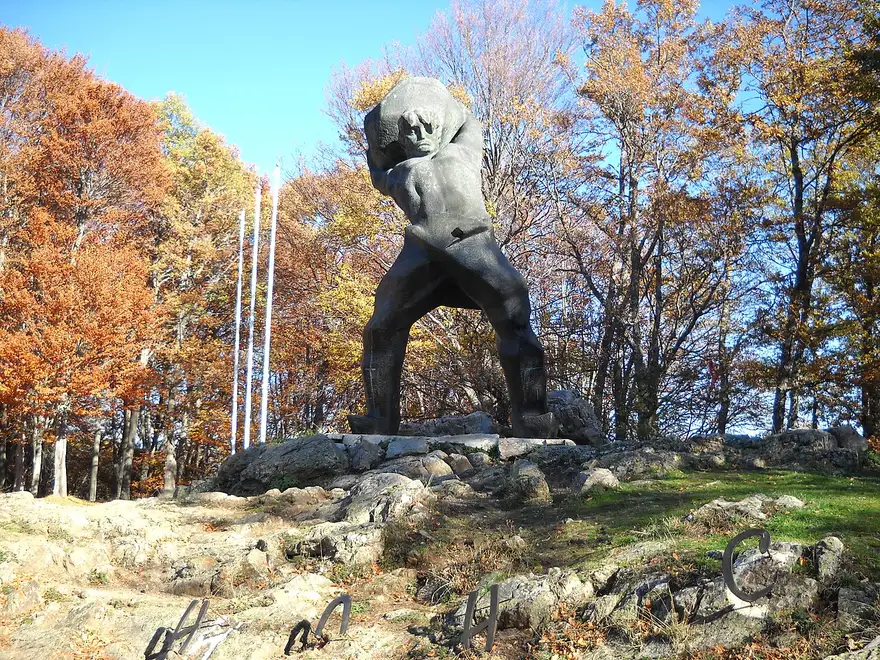 A dramatic bronze statue of a crouching fighter hoisting a boulder, the Pitu Guli monument at Mečkin Kamen near Kruševo, among autumn trees