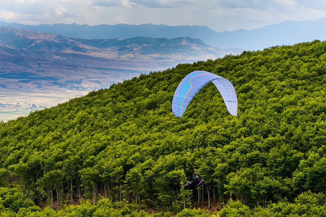 A paraglider soaring over green wooded mountain slopes above Kruševo, with the Pelagonia plain and distant mountains below