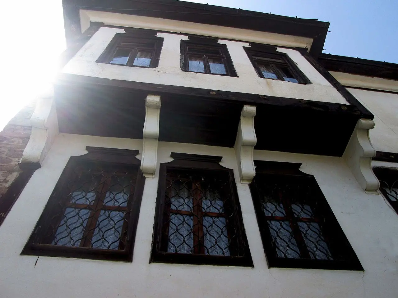 Looking up at a white-plastered Kruševo house with a jettied dark-timber upper floor and rows of latticed wooden windows