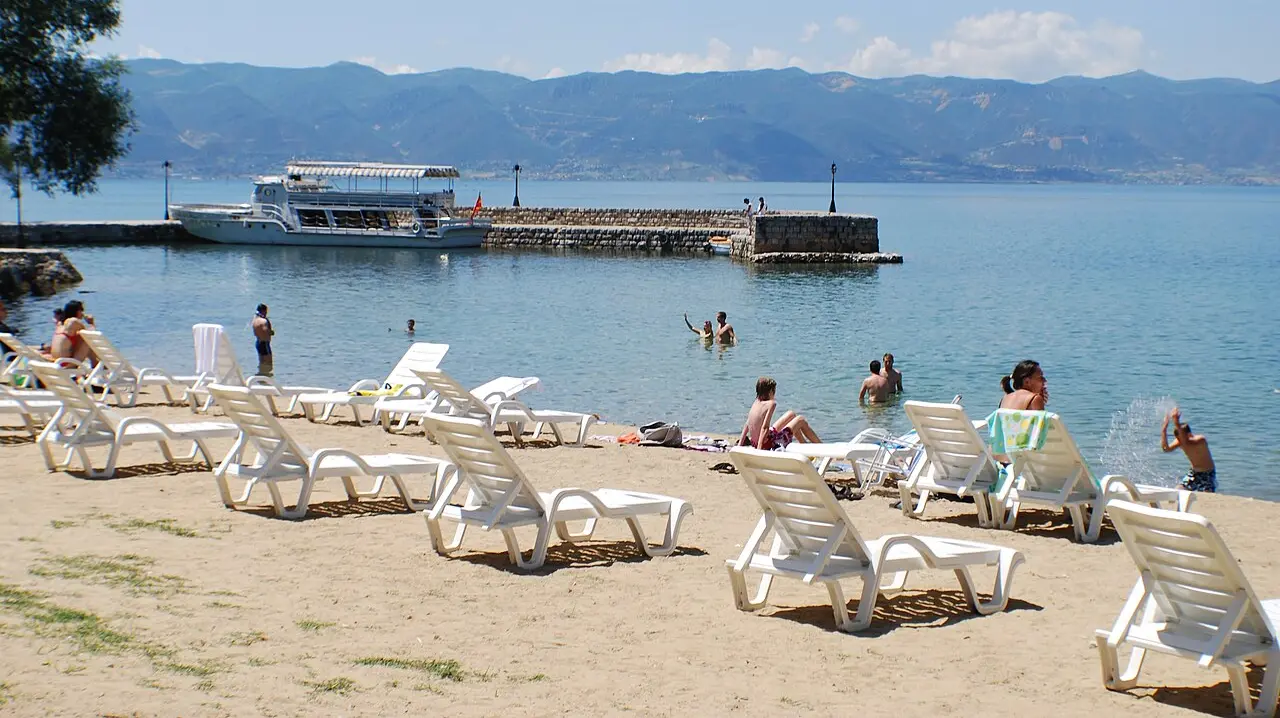 Sunbeds on a sandy beach near St. Naum on Lake Ohrid, with swimmers in clear water, a tour boat at a stone jetty and mountains behind