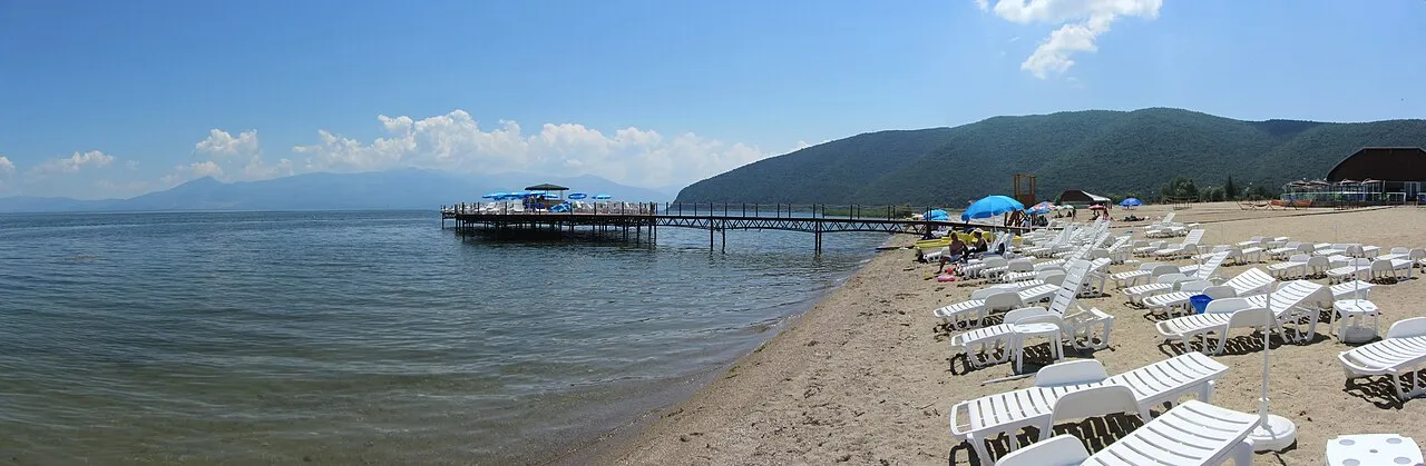 A wide sandy beach at Stenje on Lake Prespa with rows of white sunloungers, a wooden pier and green mountains across the water