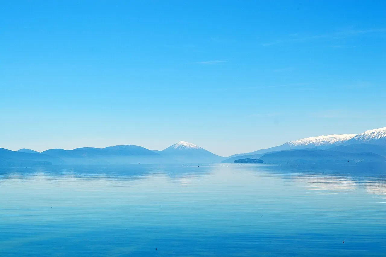 The calm blue expanse of Lake Prespa at dawn, with snow-streaked mountains and a distant island rising from the still water