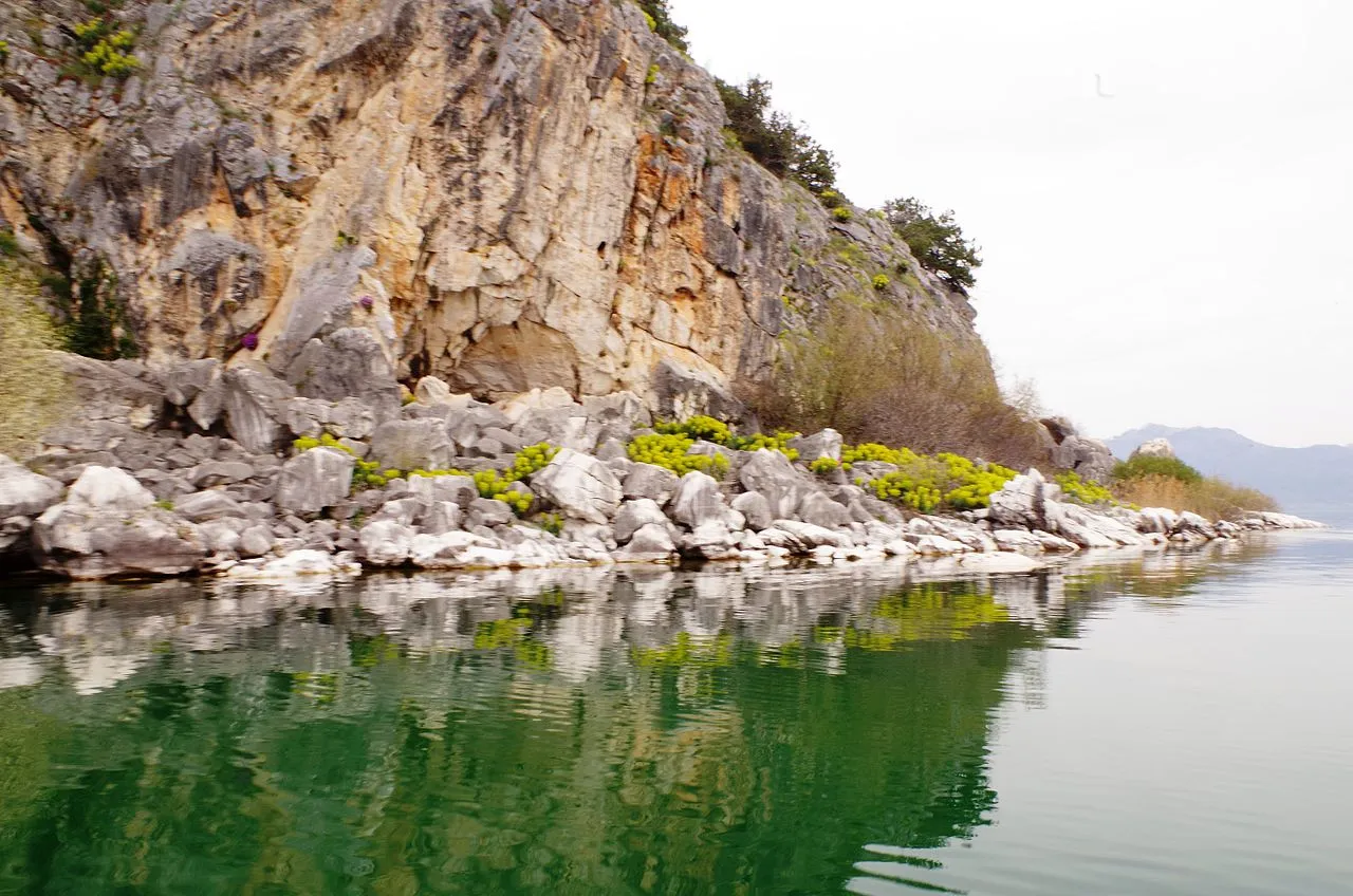 The pale limestone cliffs and boulder shoreline of Golem Grad reflected in the clear green water of Lake Prespa
