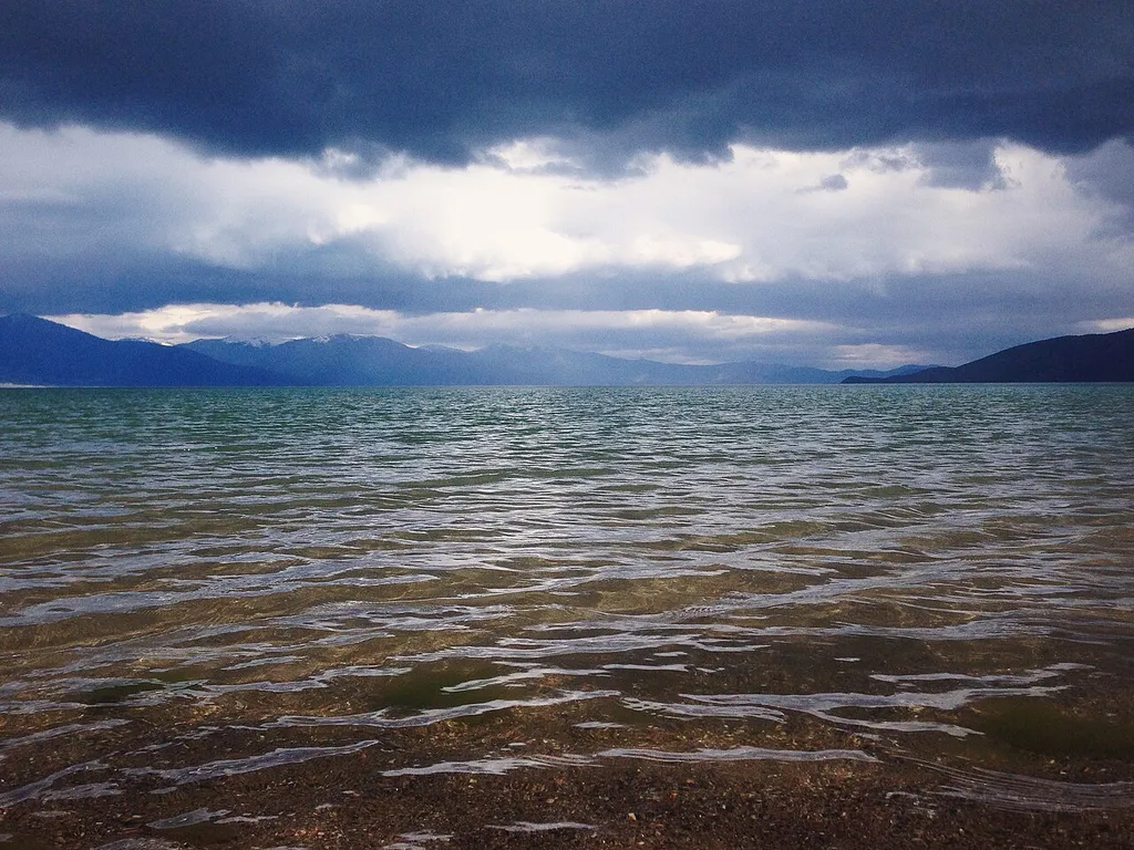 Clear shallow water over a pebble beach on Lake Prespa, with a dramatic sky and mountains across the water