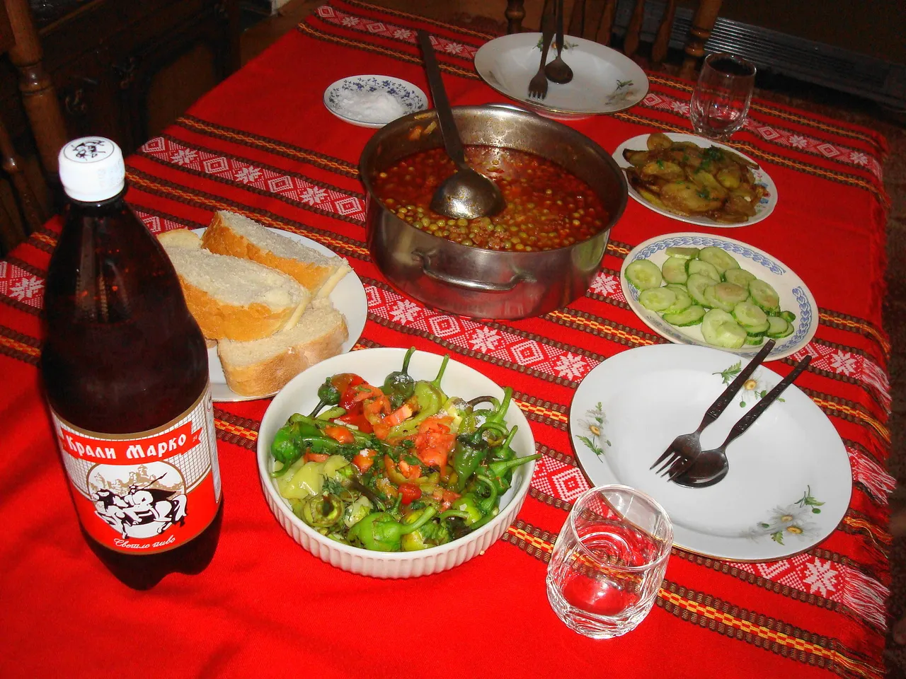 A Macedonian home table with a pot of bean stew, bread, pepper salad and a bottle of local beer on a red woven runner