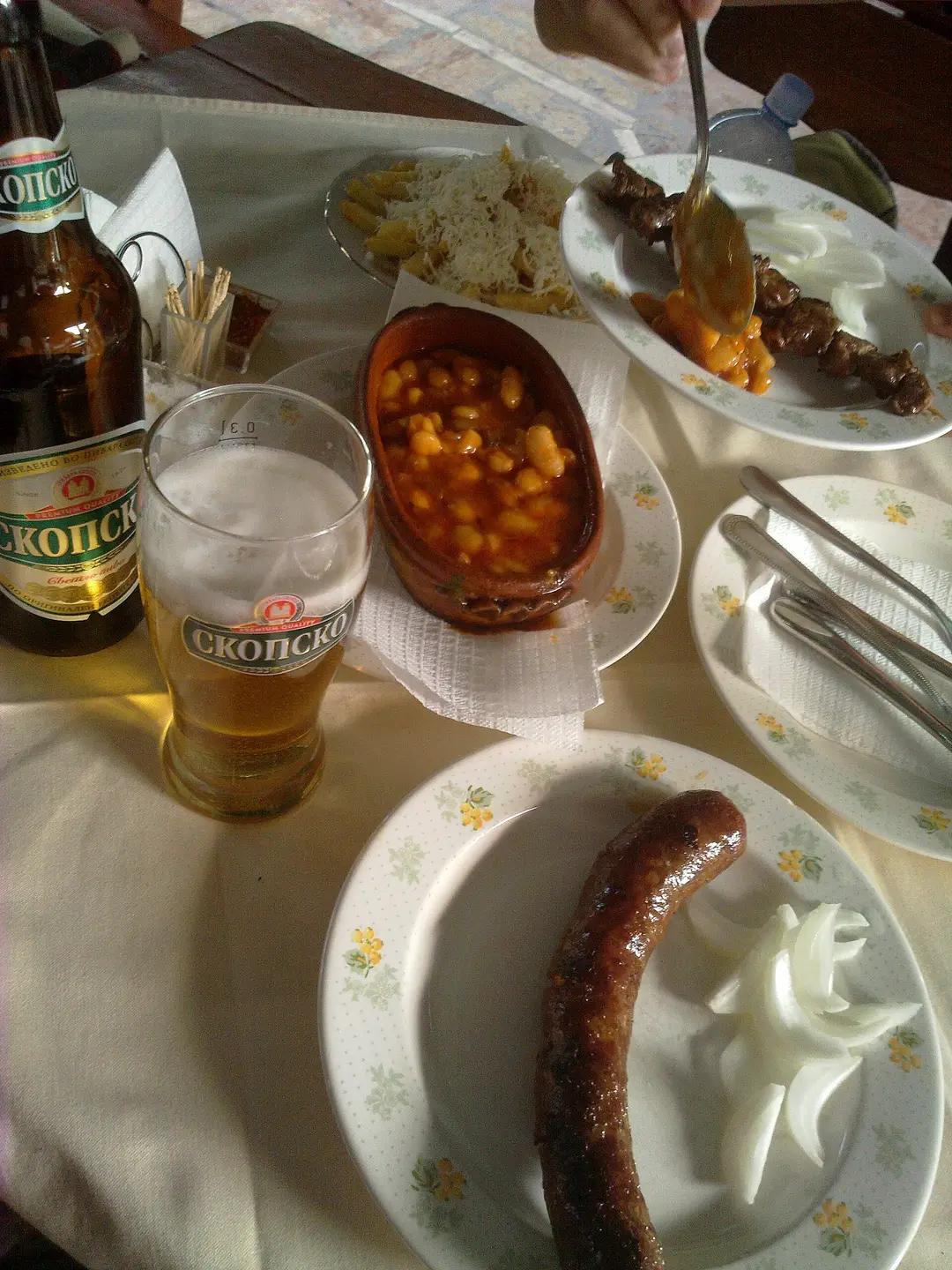 A kafana table with Skopsko beer, tavče gravče in a clay pot, a grilled skewer, sausage and onion