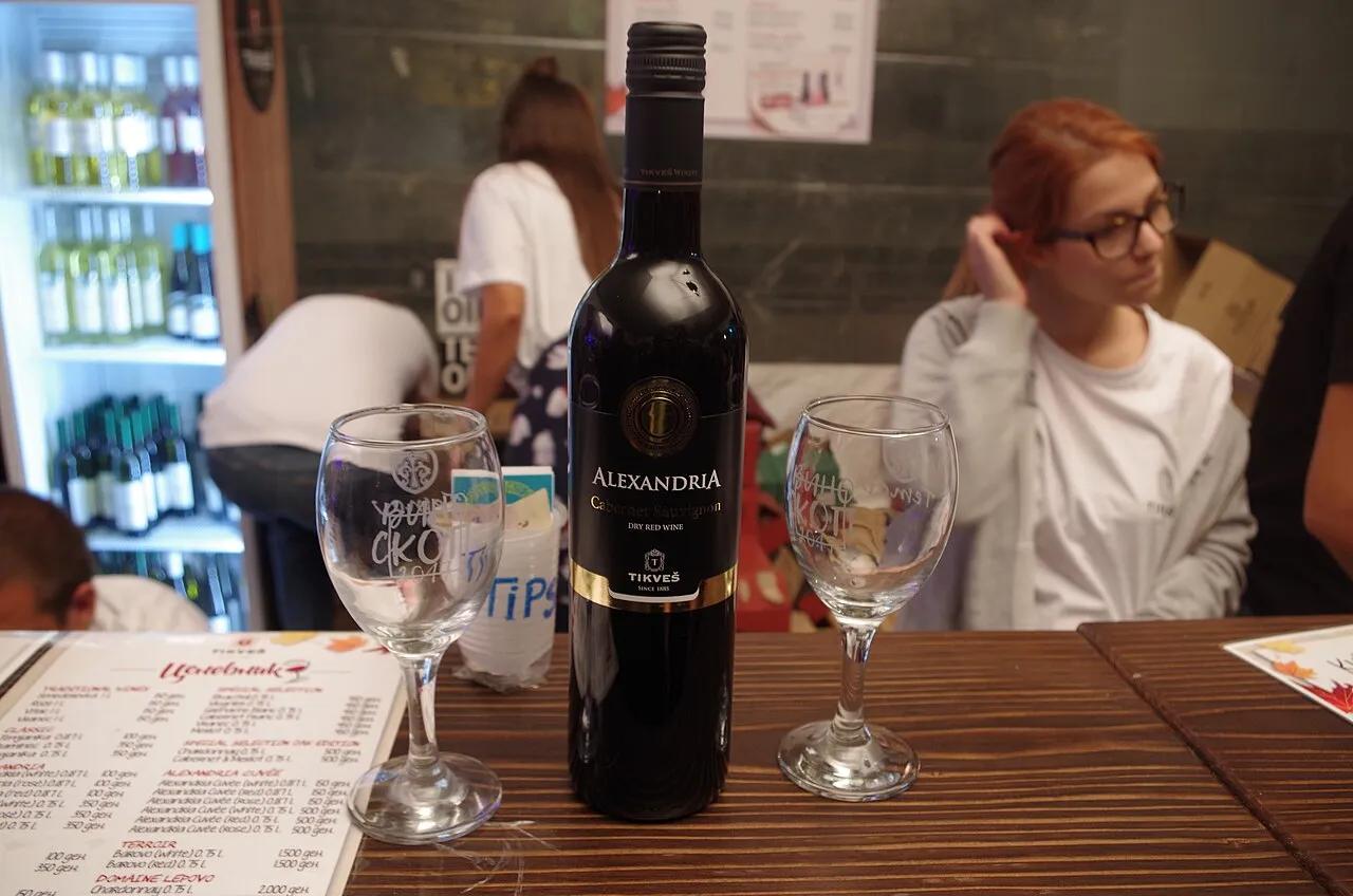 A bottle of Tikveš Alexandria dry red wine with two empty tasting glasses and a printed price list on a bar