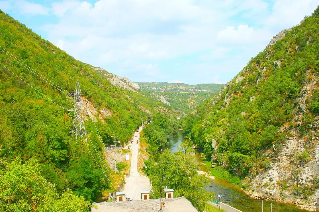 A road and walking path running along the Matka reservoir, with small boats moored below steep canyon walls