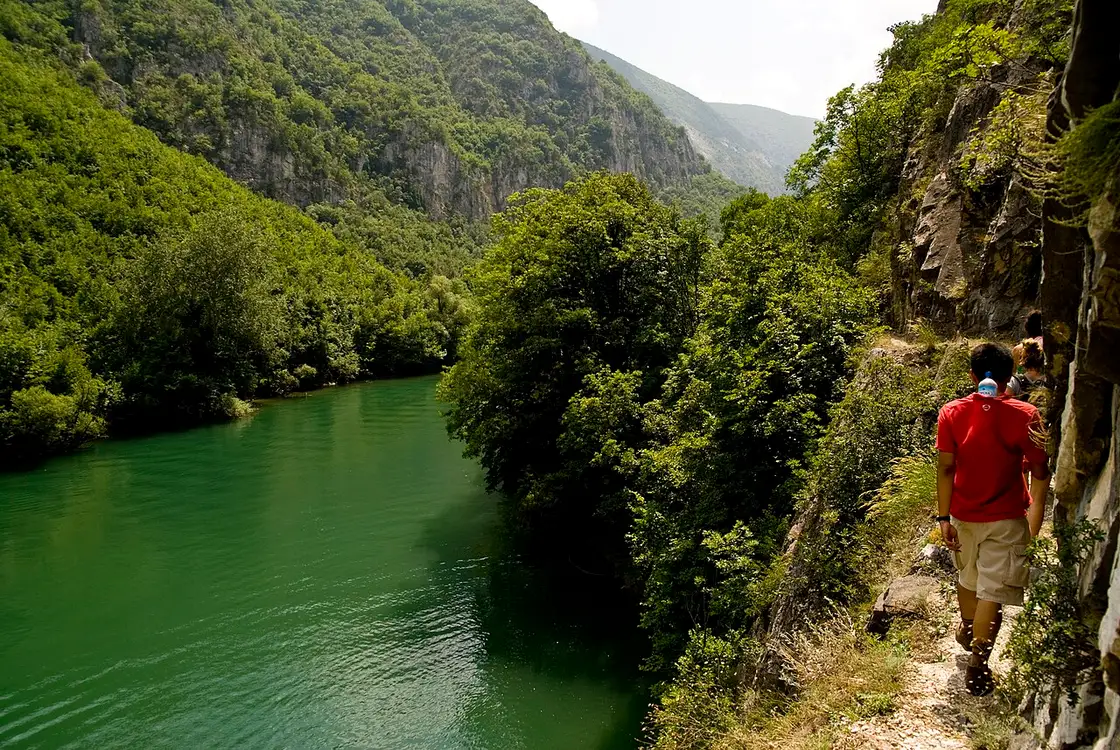 A hiker walking a narrow path along the green river inside Matka Canyon, cliffs rising on one side