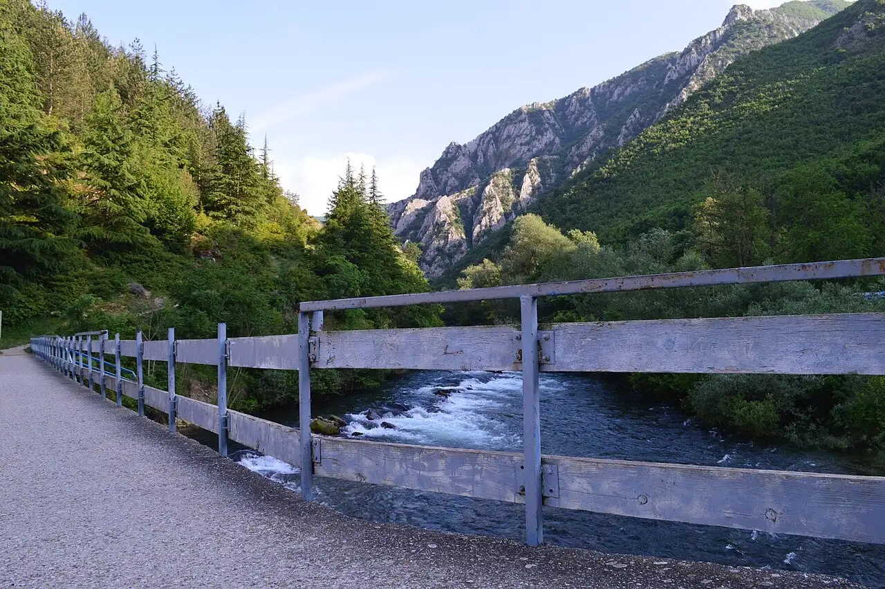 A riverside path and fence with the wooded canyon walls of Matka rising behind